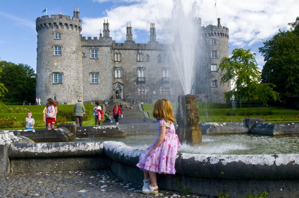 Guests enjoying a sunny day at a historic castle with a beautiful fountain, lush gardens, and family-friendly atmosphere.