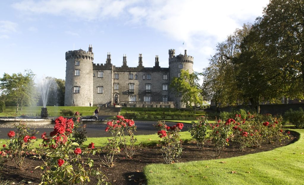 Historic castle hotel with lush gardens, elegant fountains, and vibrant roses under a bright sky, offering a serene retreat.