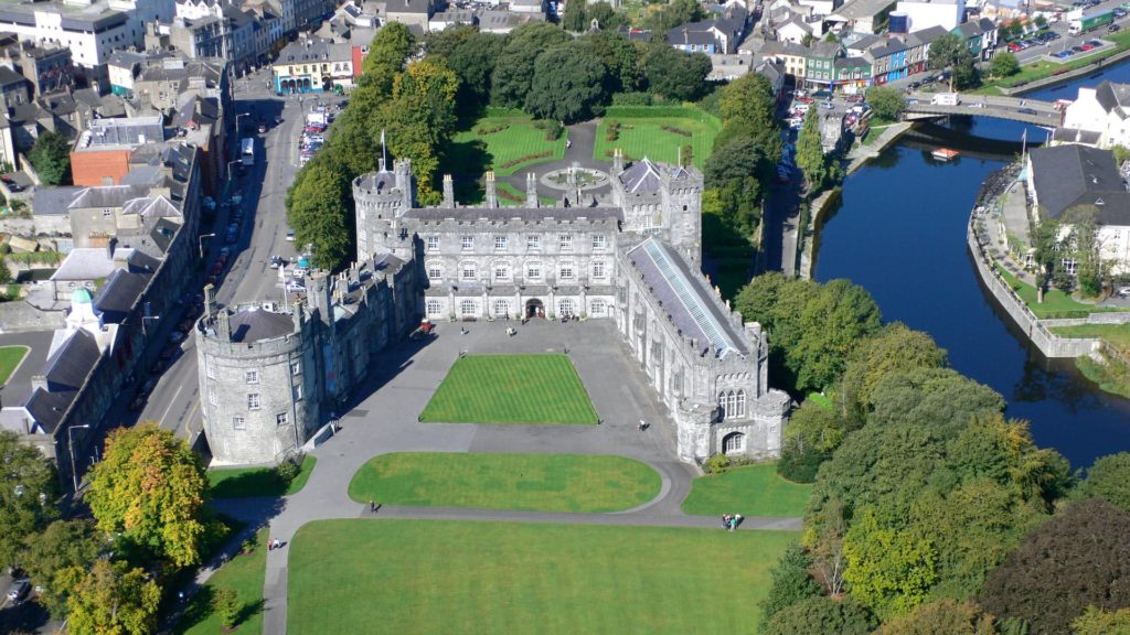 Stunning aerial view of a historic castle hotel with lush gardens, nestled by a tranquil river in a charming town.