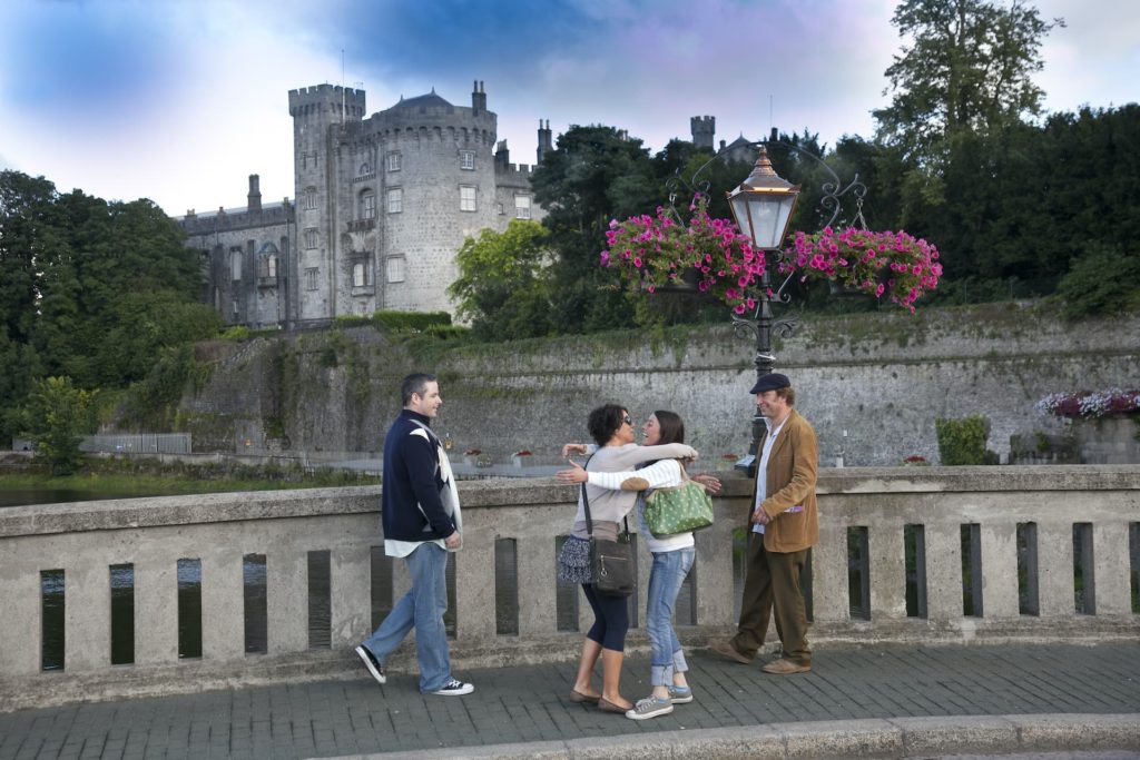 Guests enjoy a scenic walk by a historic castle, surrounded by lush greenery and vibrant flowers under a clear sky.