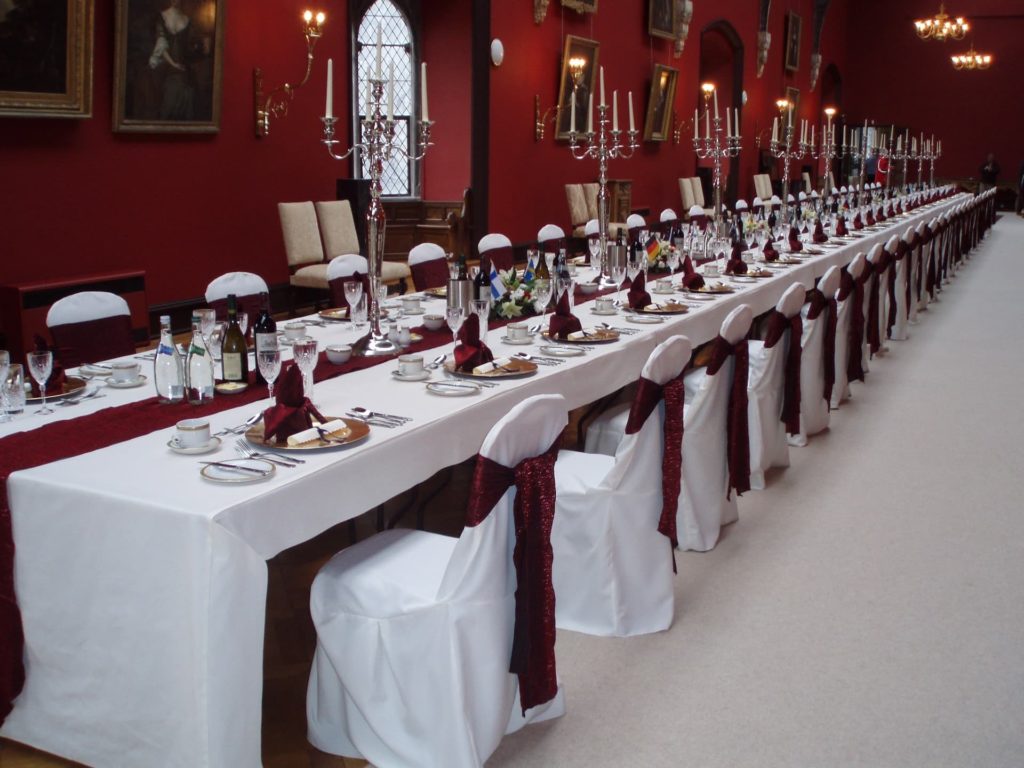 Elegant banquet hall with long dining table, red décor, white linen chairs, chandeliers, ideal for events or weddings.