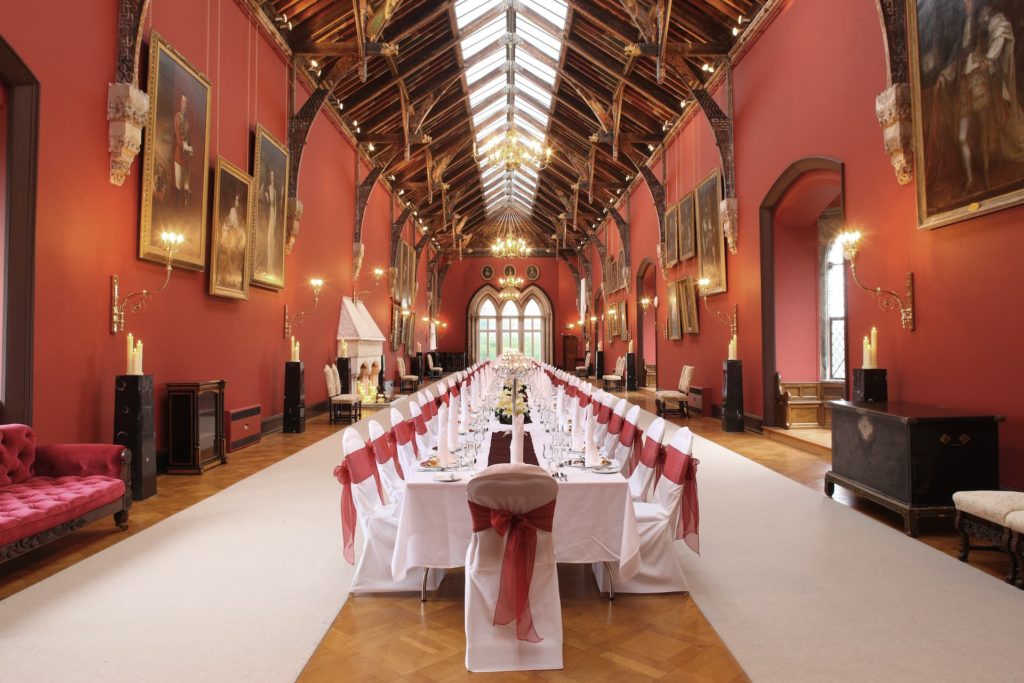 Grand dining hall with elegant red walls, ornate chandeliers, large paintings, and a long banquet table set for a luxurious event.