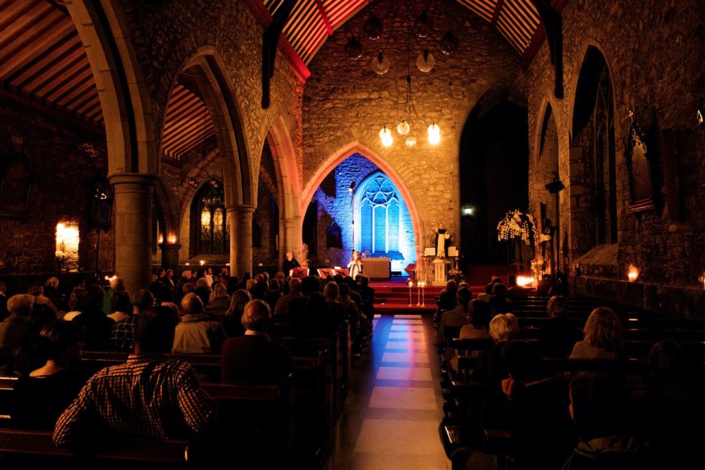 Guests enjoy a candlelit concert in a stunning stone chapel with arched ceilings and soft lighting, creating a serene atmosphere.