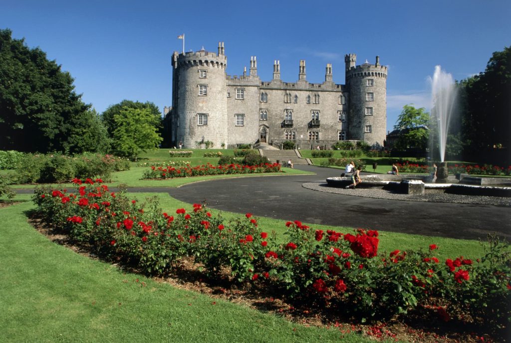 Guests relax by the elegant fountain in front of a grand, historic castle surrounded by vibrant rose gardens and lush greenery.