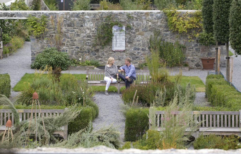 Guests relax on a bench in a serene garden surrounded by lush greenery and stone walls, offering a peaceful hotel retreat.