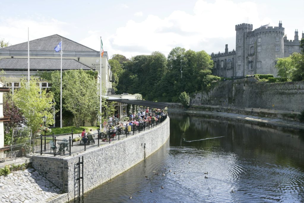 Riverside terrace with outdoor seating overlooking scenic river and castle, perfect for relaxing and enjoying nature.