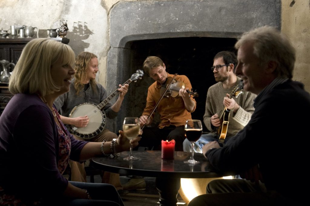 Guests enjoying live music by a cosy fireplace, with drinks on the table, creating a warm, inviting atmosphere.