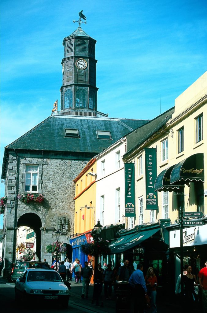 Historic clock tower over vibrant street with colourful shops, ideal for a charming city stay near attractions.