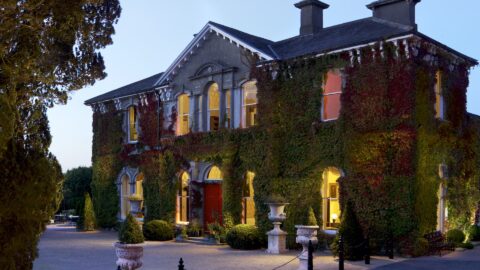 Elegant ivy-covered hotel facade with glowing windows, welcoming entrance, and serene outdoor seating at dusk.
