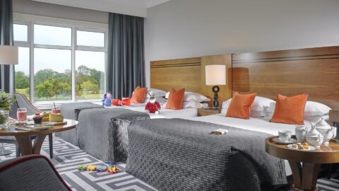 Spacious room with two luxe double beds, vibrant orange cushions, scenic window view, cosy seating and family-friendly amenities.