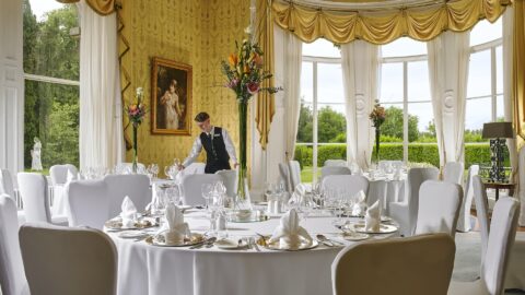 Elegant hotel dining room with large windows, lush garden views, round tables with white tablecloths, and floral centrepieces.