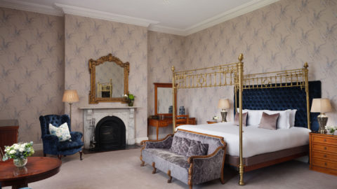 Elegant hotel room with a four-poster bed, luxurious blue velvet chairs, ornate fireplace, and classic decor. Relaxing retreat.
