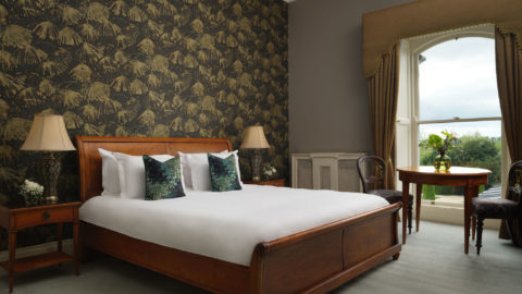 Elegant hotel room with king bed, plush pillows, nature-themed wallpaper, window view, and classic wooden furniture.