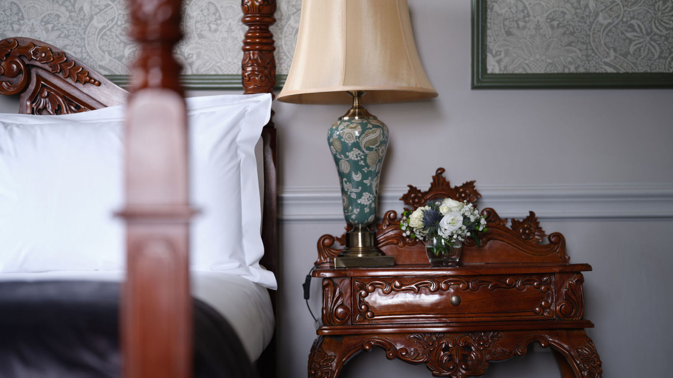 Elegant hotel room with ornate wooden bed, luxurious bedding, decorative nightstand, classic lamp, and floral arrangement.