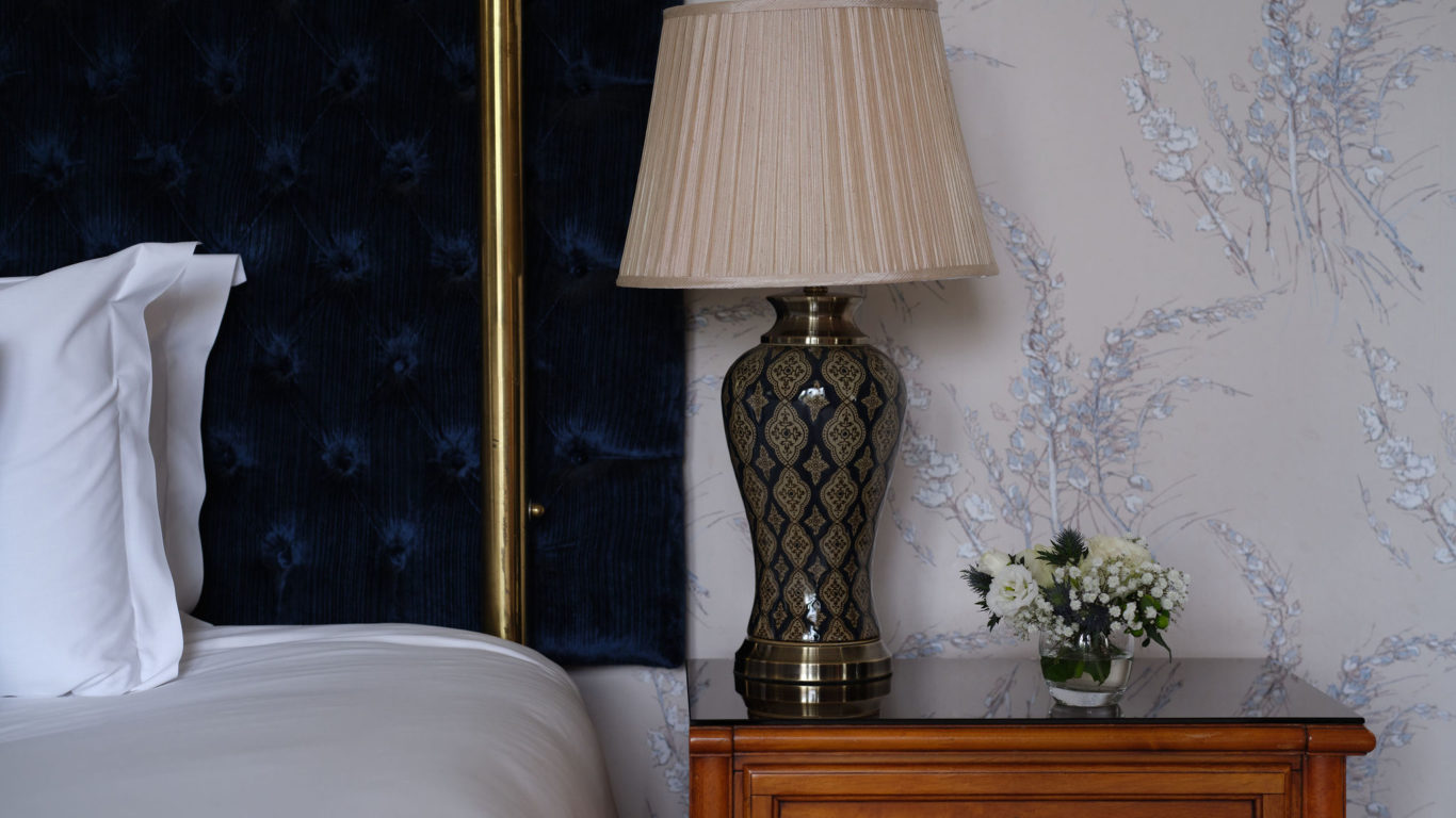 Elegant hotel room with a plush blue headboard, ornate lamp, and fresh flowers on a wooden bedside table. Relaxing ambiance.