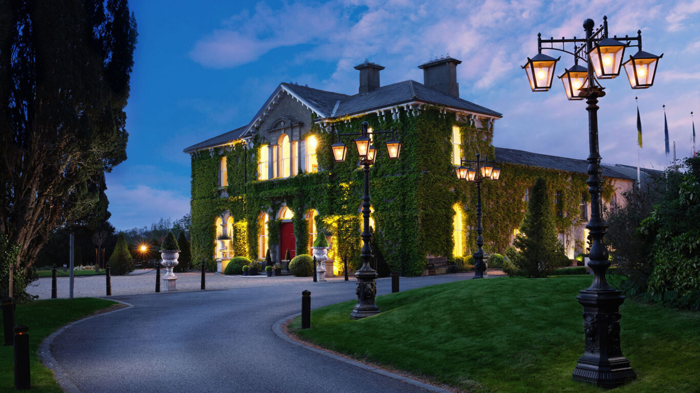 Charming ivy-covered hotel facade at dusk, warmly lit windows, elegant lamp posts along a winding driveway.