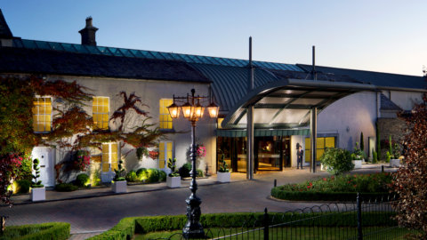 Elegant hotel entrance at dusk, with warm lighting, lush gardens, classic architecture, welcoming ambiance.