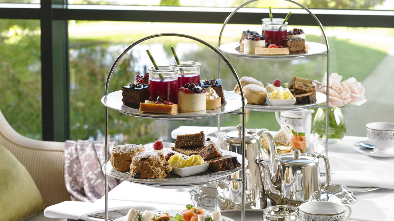 Three-tiered trays with assorted pastries, cakes, and sandwiches on a table set for afternoon tea with teapots, cups, and saucers in front of a large window.