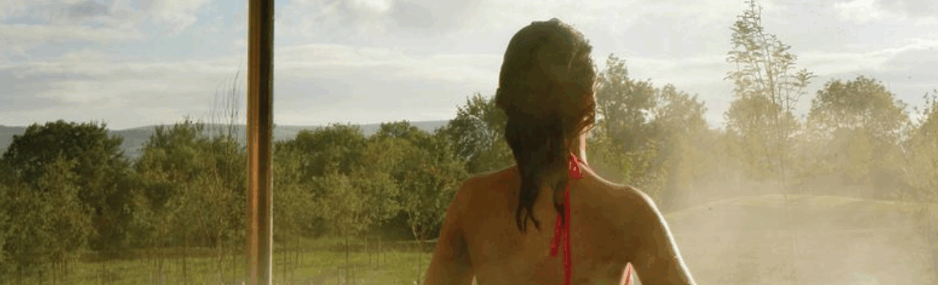 A person with long hair in a red swimsuit sits in an outdoor hot tub, facing a misty landscape with trees and cloudy sky.