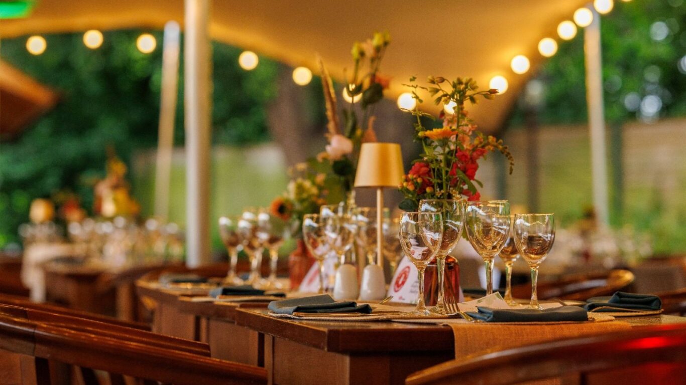 A decorated outdoor dining area with wooden tables set with glassware, napkins, and floral centerpieces under string lights.