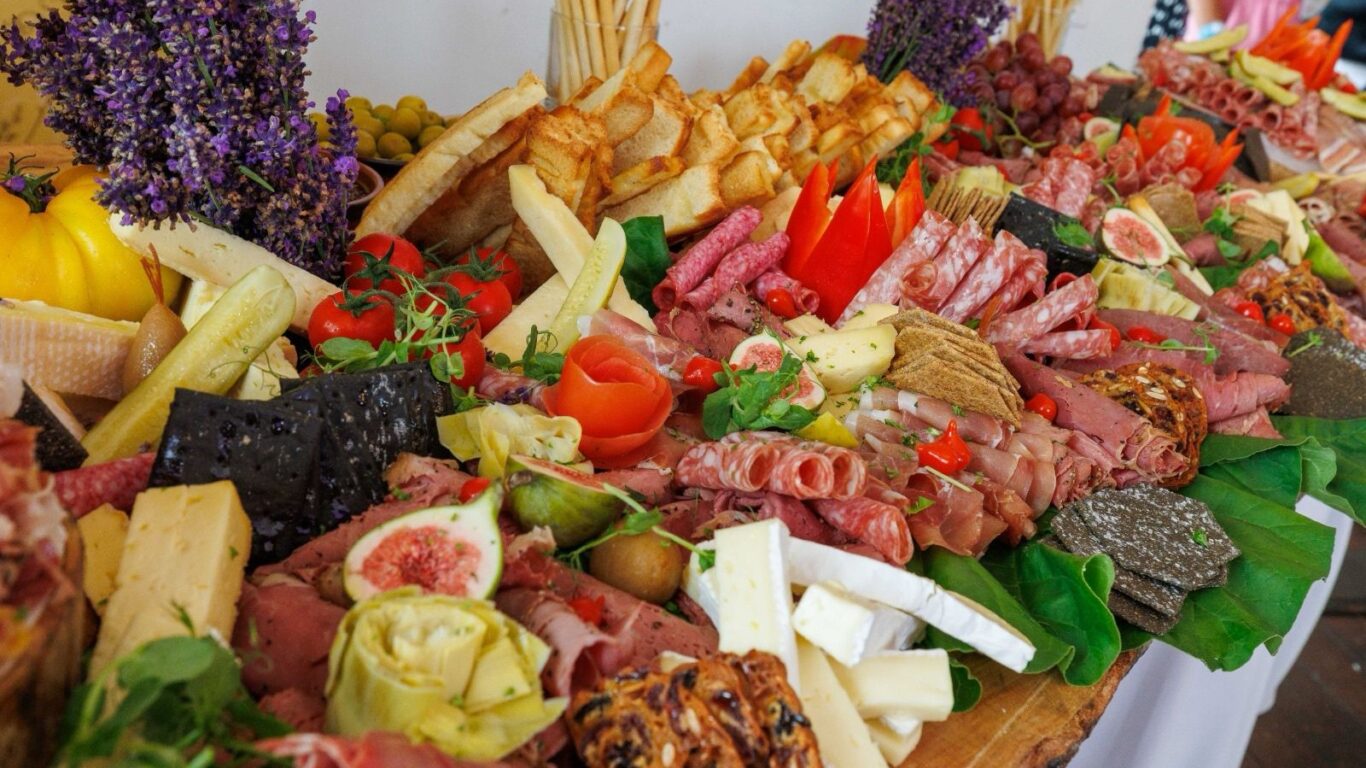 A large charcuterie board with assorted cheeses, cured meats, crackers, fresh figs, bread, olives, artichokes, and herbs, arranged on a wooden surface with purple flowers in the background.