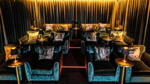 A small home theater with plush teal armchairs, gold side tables with black lamps, and heavy curtains covering the walls, all softly lit with warm lighting.
