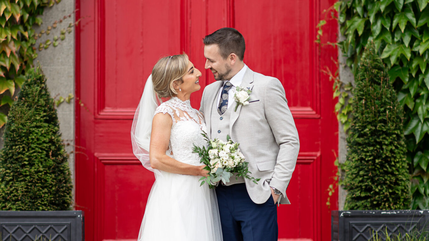 A bride and groom stand together smiling in front of a large red door, surrounded by green plants and wearing formal wedding attire.