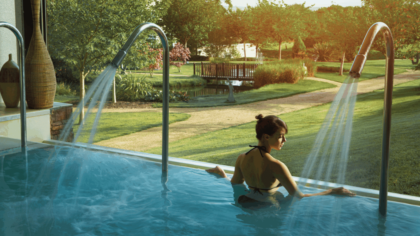Guest relaxes in indoor pool overlooking serene gardens and fountain, soaking up morning sunlight at luxury hotel spa.