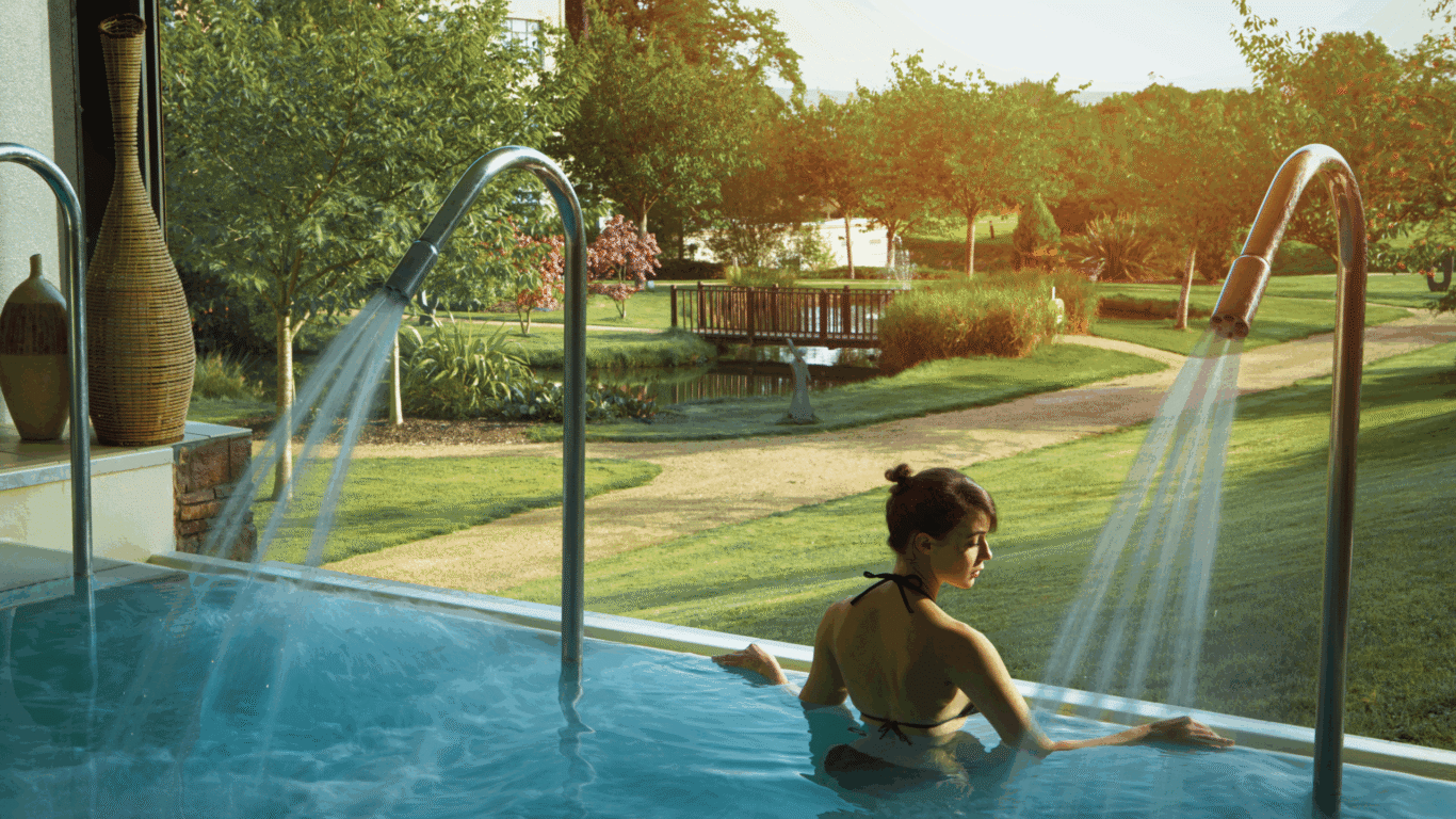 Guest relaxes in indoor pool overlooking serene gardens and fountain, soaking up morning sunlight at luxury hotel spa.