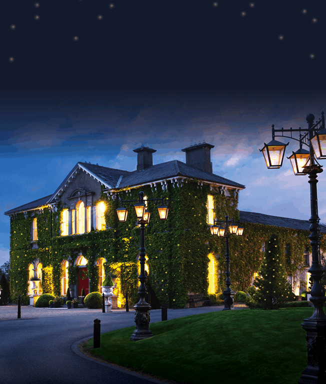 A large ivy-covered building is illuminated at night, with glowing windows, decorative street lamps, and a grassy area with a lit tree in front.