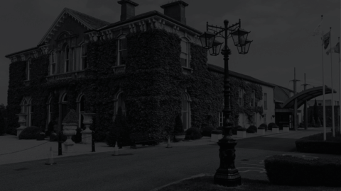 A large, ivy-covered brick building with chimneys and arched windows stands beside a driveway and a vintage streetlamp; flags are visible near the entrance.
