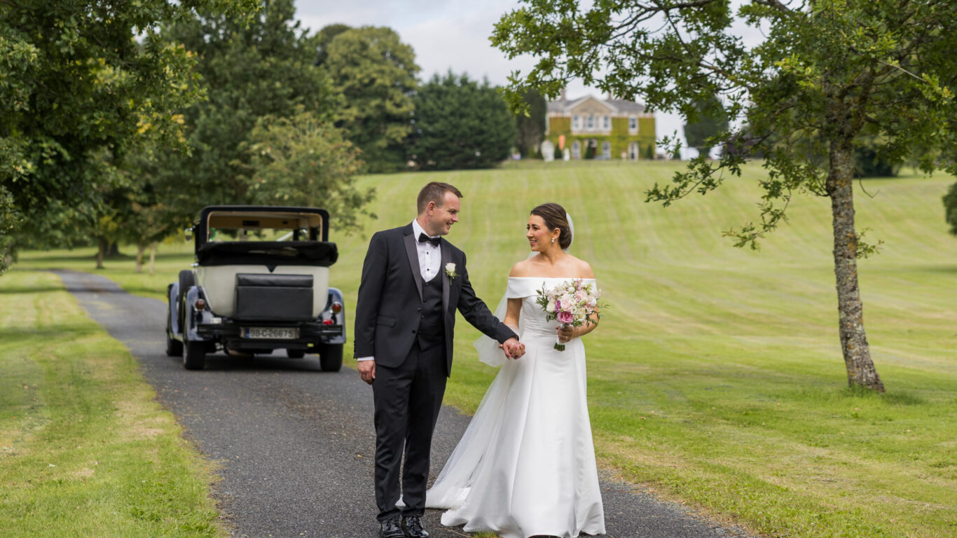 A bride and groom walk hand in hand on a driveway lined with grass and trees, with a vintage car and a large house in the background.