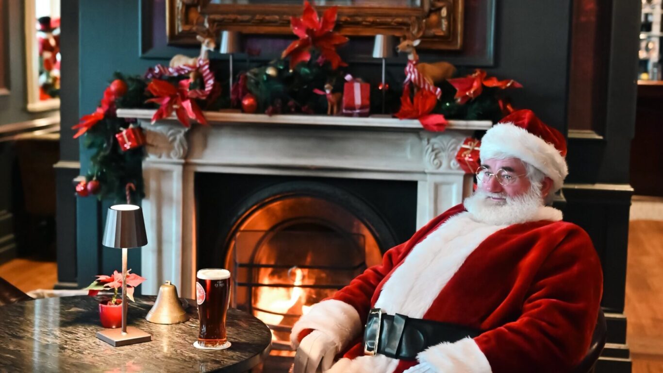 A person dressed as Santa Claus sits in an armchair by a lit fireplace in a decorated room, with drinks on the table nearby.