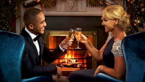 A man and woman dressed formally clink champagne glasses while sitting by a lit fireplace with festive decorations in the background.