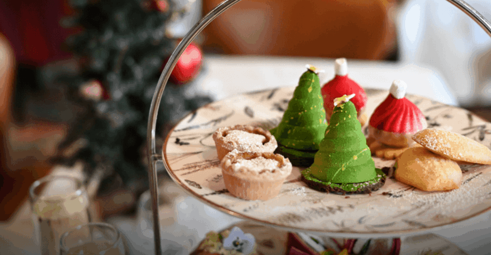 A plate of assorted festive pastries, including tree-shaped green desserts, red confections with white tops, and powdered mini tarts, is displayed on a tiered stand with a blurred Christmas tree in the background.