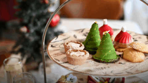 A plate of assorted festive pastries, including tree-shaped green desserts, red confections with white tops, and powdered mini tarts, is displayed on a tiered stand with a blurred Christmas tree in the background.