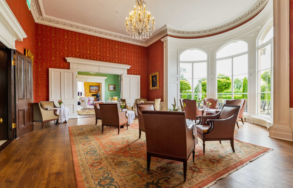 Elegant lounge with armchairs and tables on a patterned rug, large curved windows, red patterned walls, and a chandelier. Outdoor greenery visible through the windows.