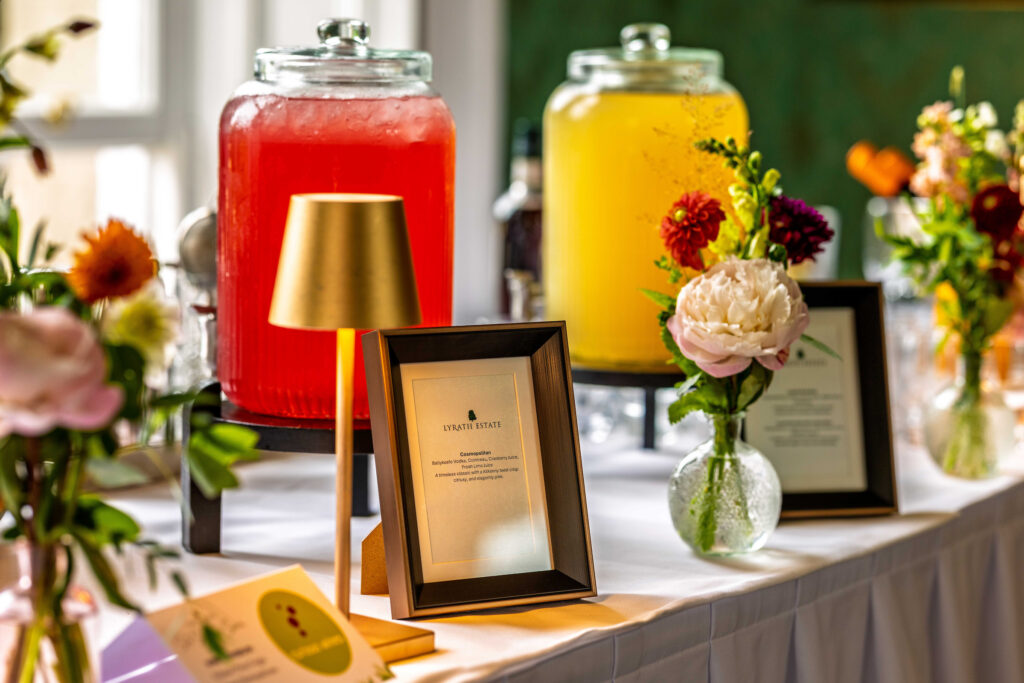 A table with two large glass dispensers of red and yellow drinks, framed signs, flowers in vases, and a small gold lamp.