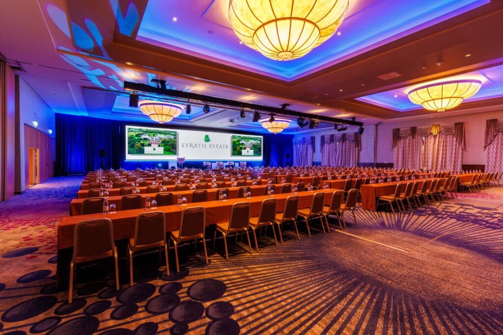 A large conference room with rows of tables and chairs facing a stage with three projection screens displaying 
