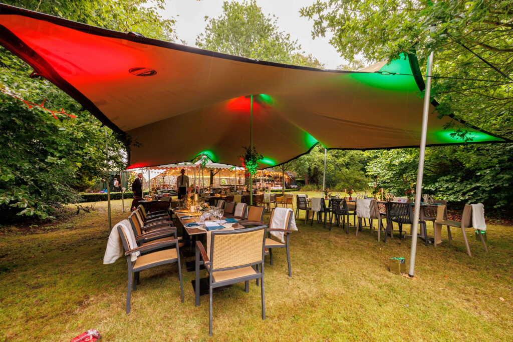 Outdoor dining area under a large canopy with red and green lights, set with tables and chairs, surrounded by trees and greenery.