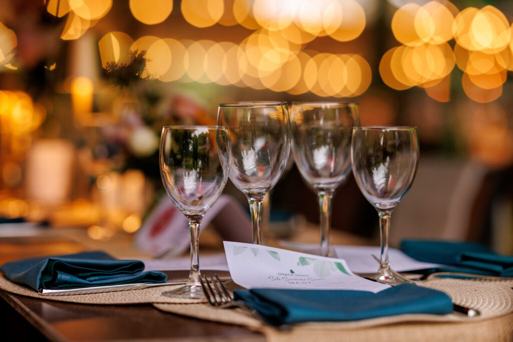 A close-up of a set dining table with four empty wine glasses, teal napkins, a menu card, and blurred warm lights in the background.