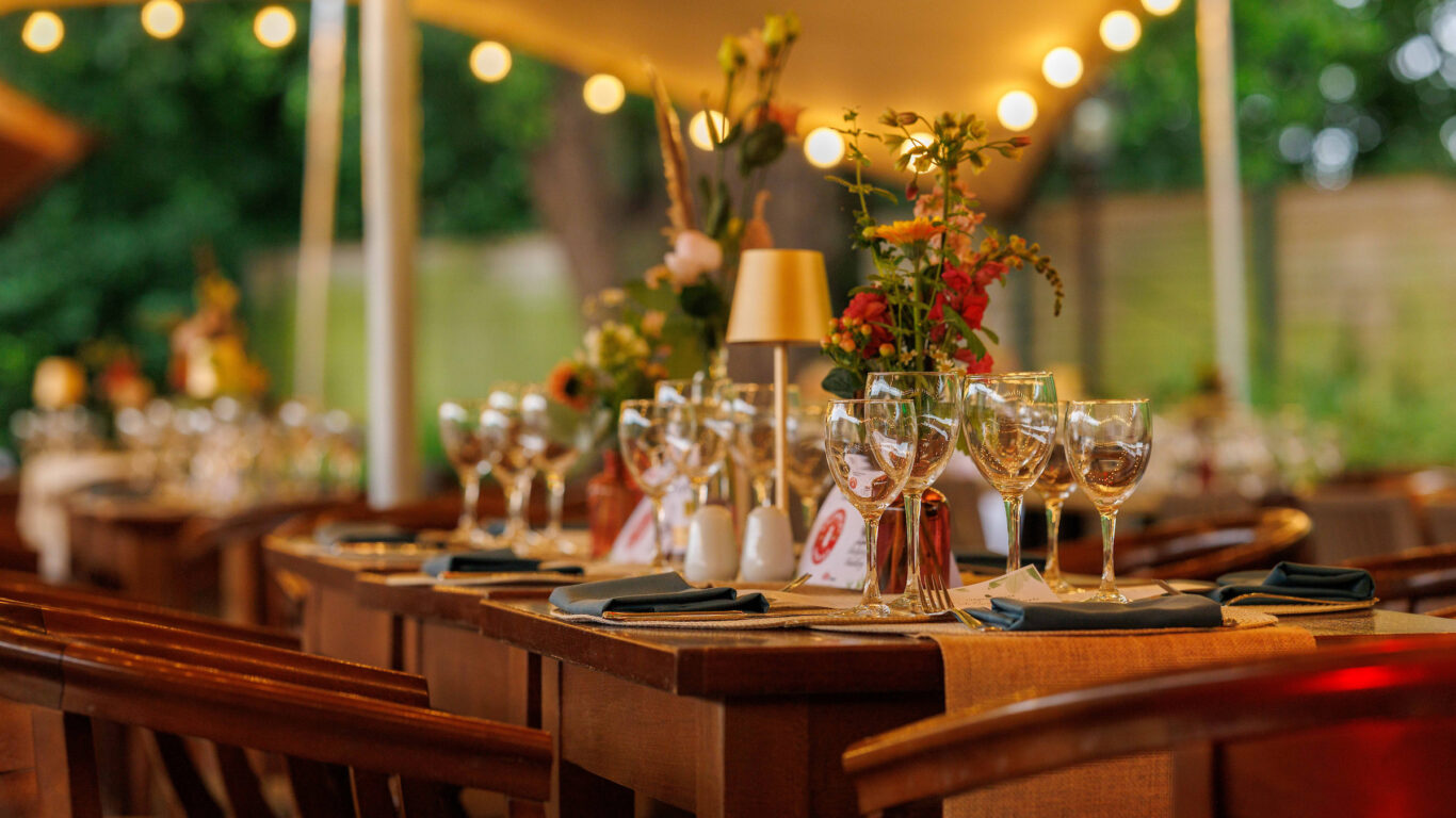 A set dining table with glasses, plates, and floral centerpieces under string lights in an outdoor setting.