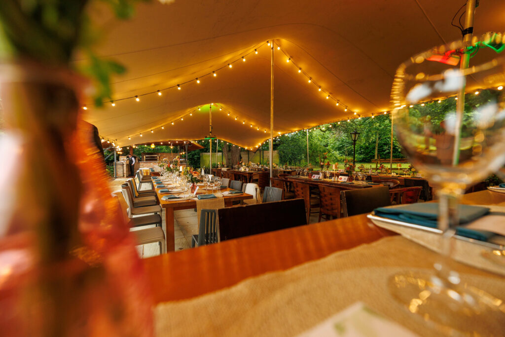 A long dining table set for an outdoor event under a large tent with string lights, surrounded by trees and additional tables.