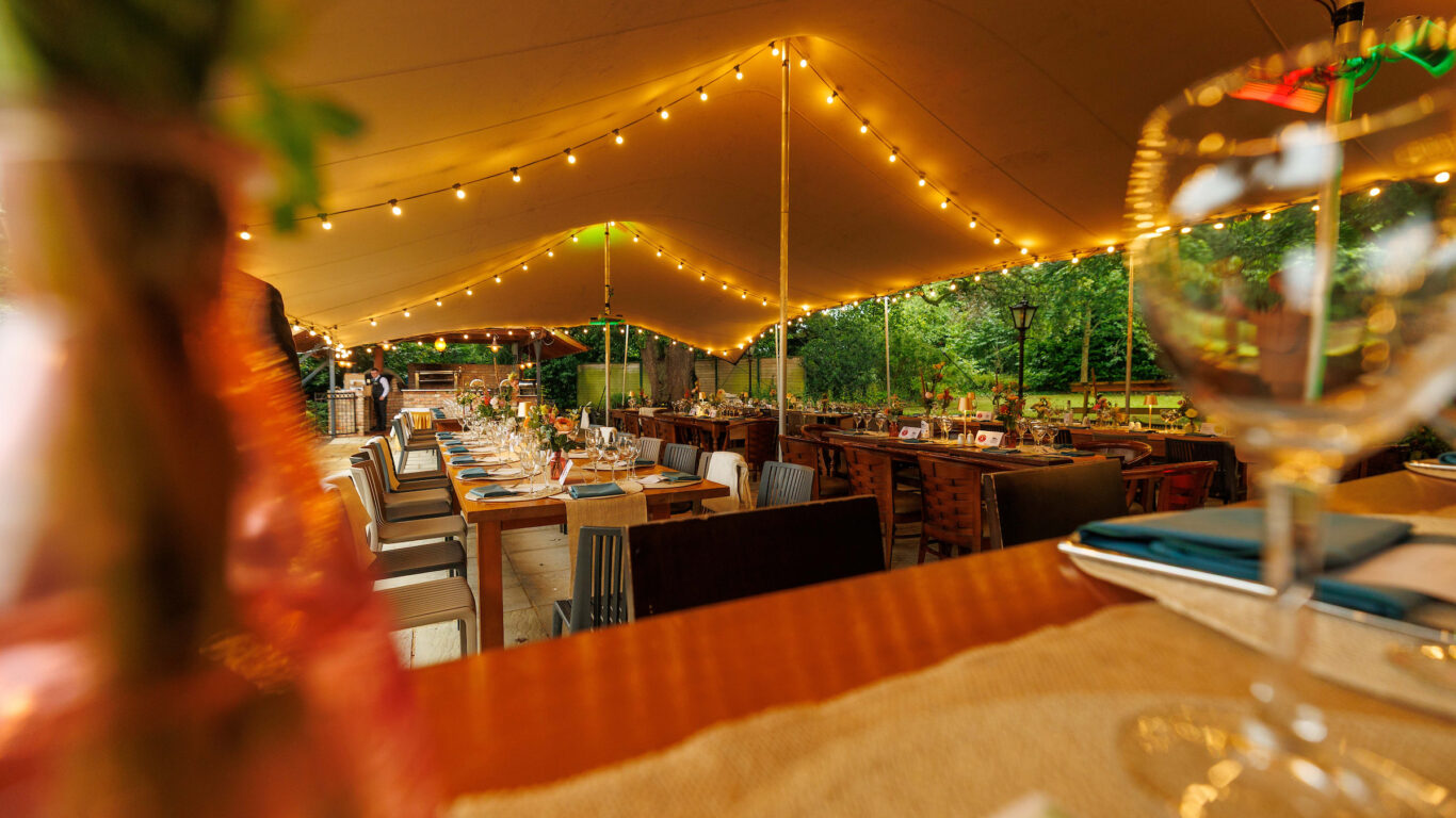 A long dining table set for an outdoor event under a large tent with string lights, surrounded by trees and additional tables.