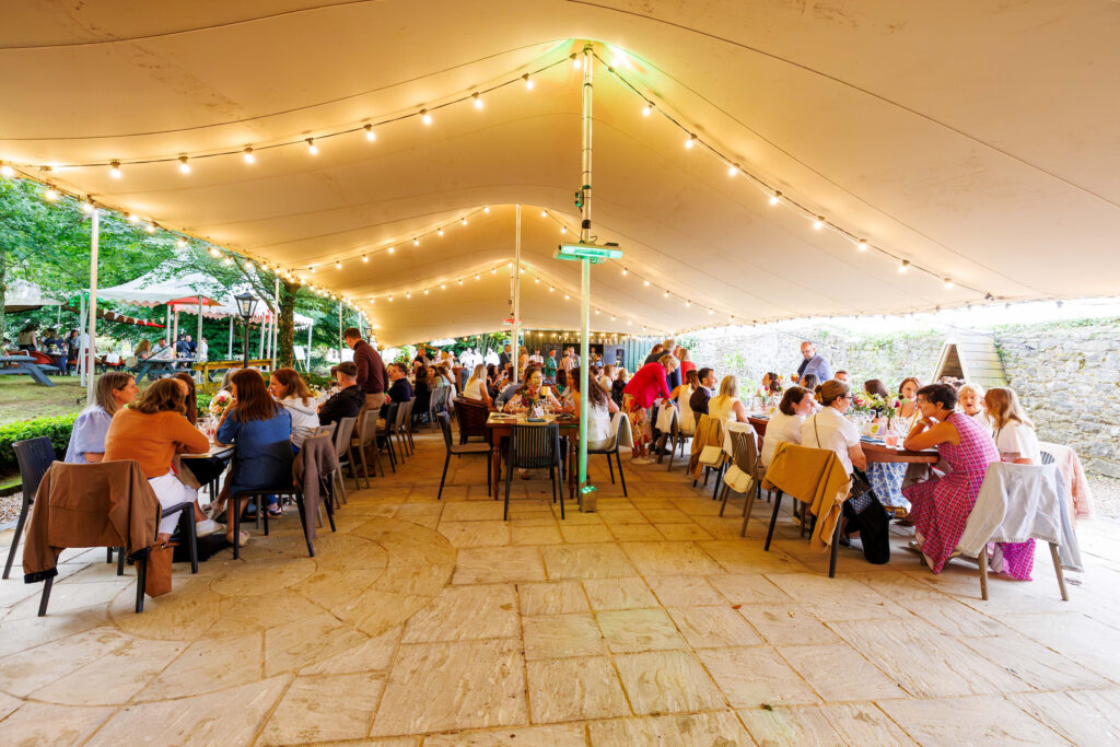 People are seated at tables under a large outdoor tent with string lights, having a meal and talking in a garden setting.