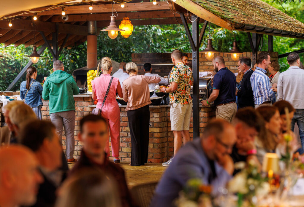 People stand in line at an outdoor buffet while others sit at tables under a covered patio decorated with lights and flowers.