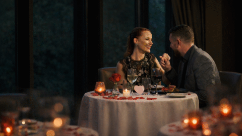 A couple sits at a candlelit restaurant table decorated with rose petals, smiling and holding hands while enjoying drinks.