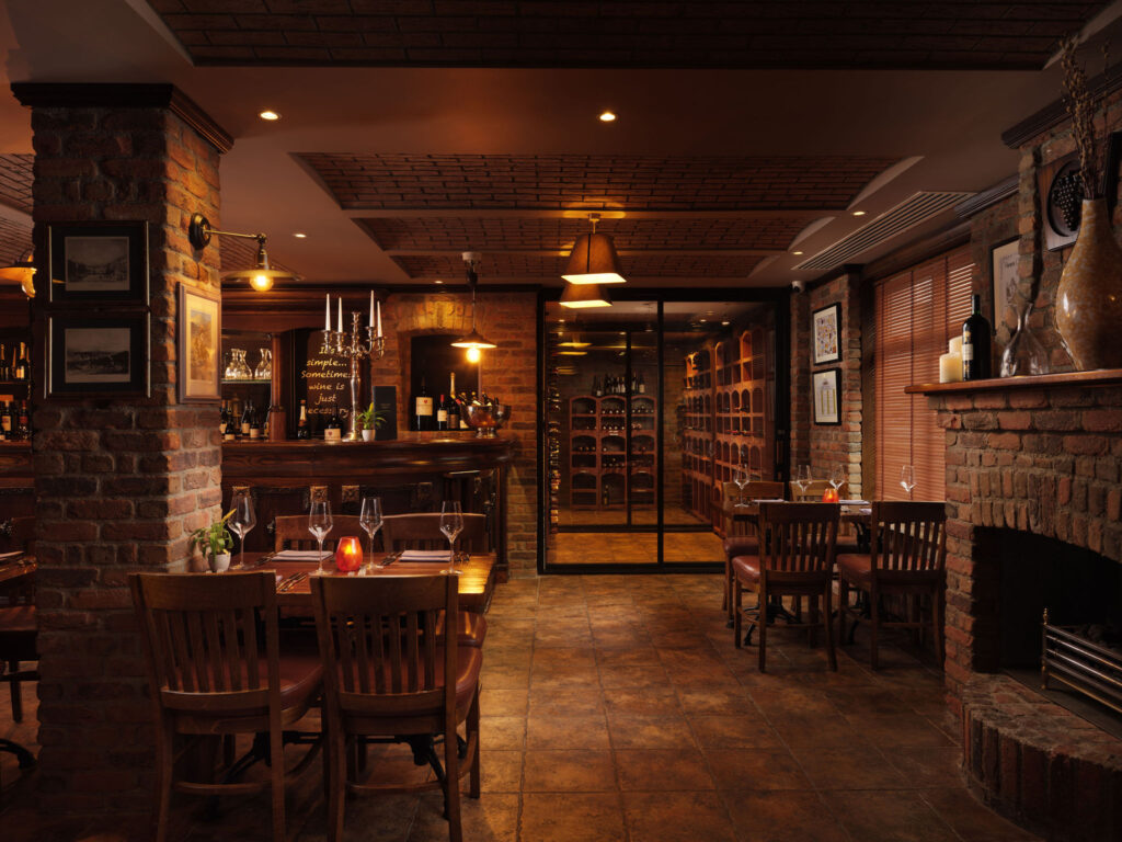 Cozy restaurant interior with brick walls, wooden tables and chairs, dim lighting, a bar counter, and a glass wine cellar in the background.