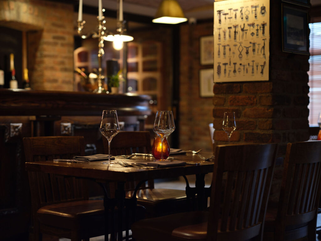 A dimly lit restaurant with a set table for four, wine glasses, a candle centerpiece, and exposed brick walls decorated with framed items and antique keys.