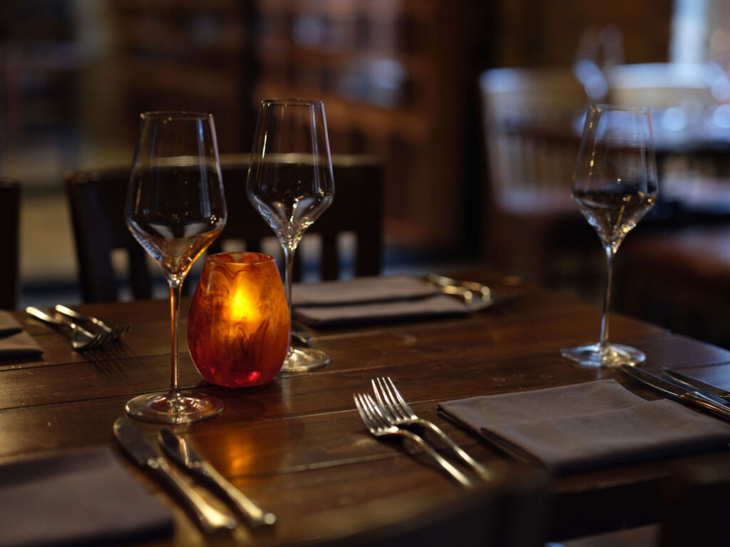 A dimly lit restaurant table set with wine glasses, cutlery, napkins, and a lit orange candle in the center.
